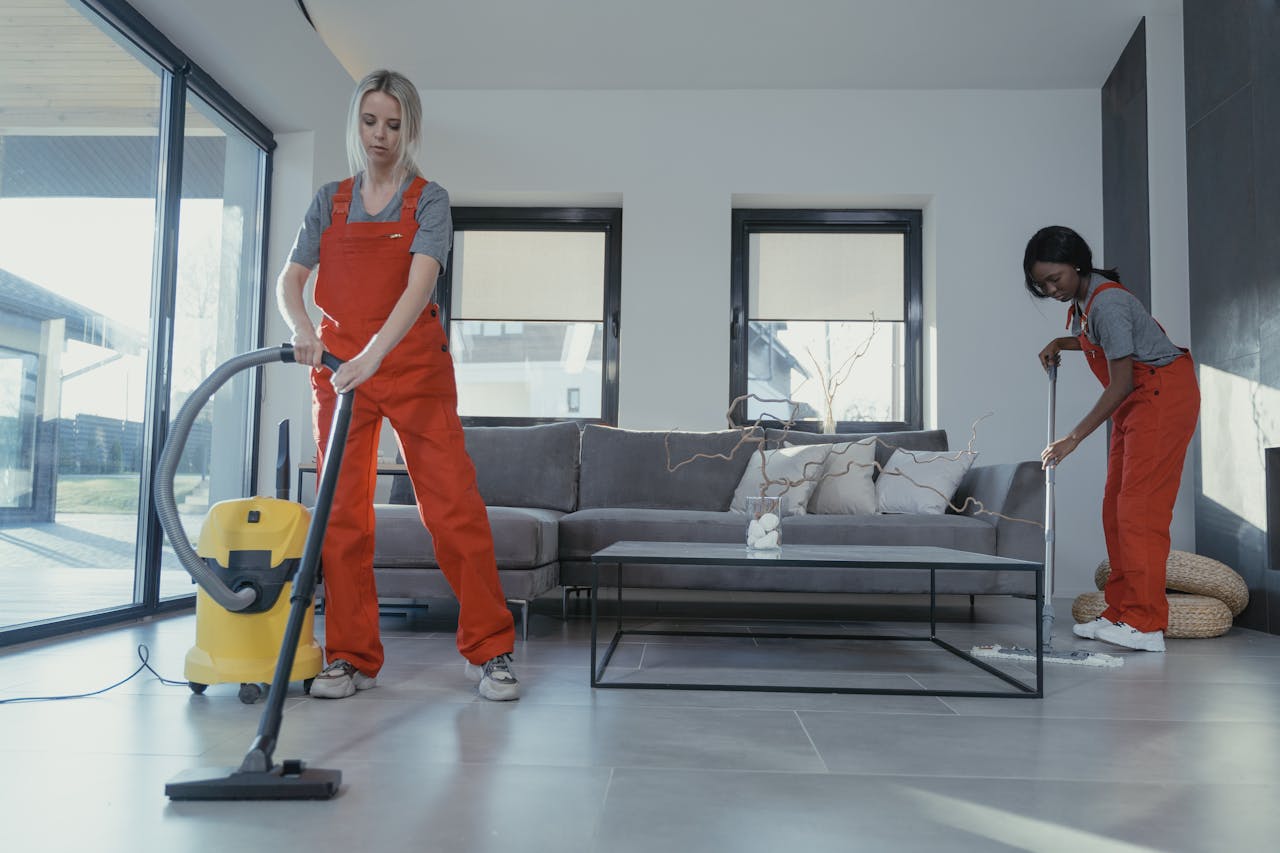 hero-img Two women in red overalls vacuuming and sweeping a modern living room.